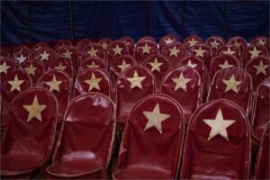 Cadeiras vermelhas vazias numa plateia de circo. No enconsto de cada cadeira tem uma estrela, menos em uma.