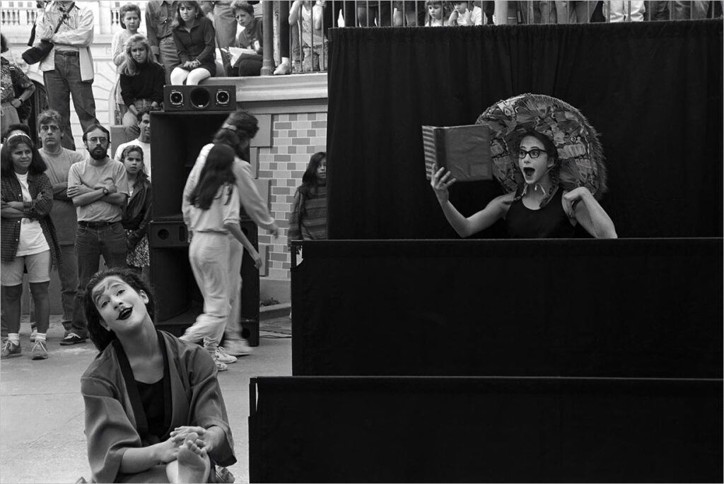 Uma foto em preto e branco de duas artistas em um palco ao ar livre. Uma, de cara pintada de branco e grandes sobrancelhas e boca preta, está sentada e agindo levemente surpresa. O outro está de pé, com um grande chapéu e blusa preta, com um livro aberto, parecendo chocada. Uma multidão os observa. Foto: Guto Muniz