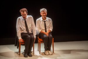 Dois homens de camisa branca e gravata preta sentam-se em cadeiras de madeira contra um fundo escuro. O homem da esquerda senta-se com a boca aberta e seus óculos, enquanto o homem da direita senta-se sorrindo gentilmente. Ambos têm posturas relaxadas. Foto: Guto Muniz