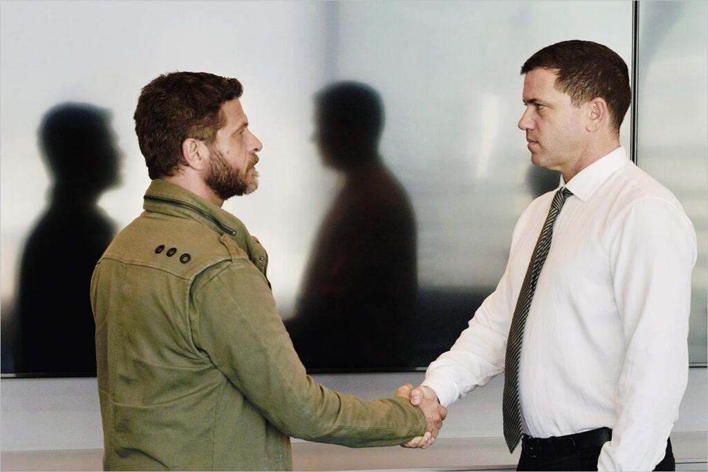 Na foto, dois homens se cumprimentam enquanto as sombras de ambos estão projetadas ao fundo sem que o cumprimento apareça. Foto: Pietro Sargentelli