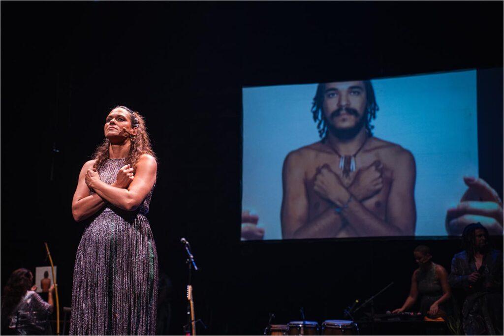 A foto mostra uma mulher em pé, de braços cruzados na forma de um X. Ao fundo, projetada em uma tela está a fotografia de um homem (Marku Ribas) que repete o mesmo gesto. Foto: Pablo Bernardo