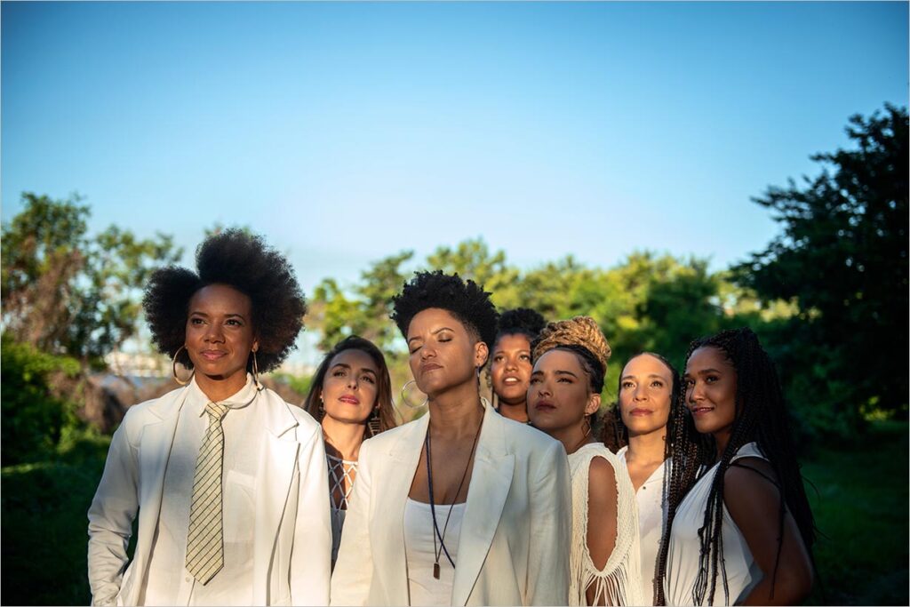 A foto mostra, da cintura para cima, sete mulheres vestidas de branco, entre luzes e sombras. Elas estão ao ar livre. Atrás delas, muitas árvores e um céu azul sem nuvens. Foto: Ira Barillo