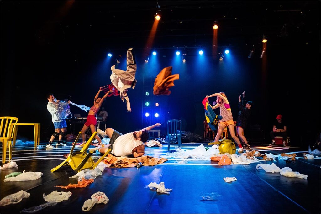 Uma cena dinâmica de palco mostra artistas jogando roupas energicamente e se movendo entre sacos plásticos e cadeiras espalhados. Luzes coloridas lançam tons vibrantes na dança energética, criando uma atmosfera caótica e animada. Foto: Carol Castanho