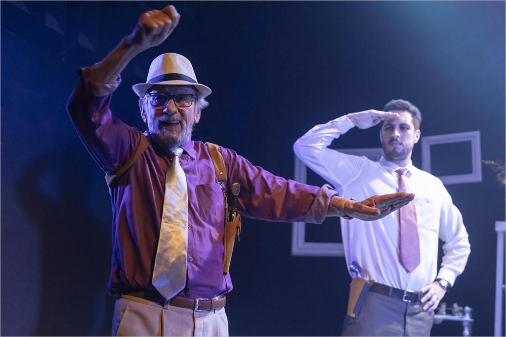 Dois homens se apresentam no palco. O homem mais velho, de chapéu e suspensórios, faz uma pose brincalhona, enquanto o homem mais jovem, de gravata, fica atrás, batendo continência. Foto: João Caldas