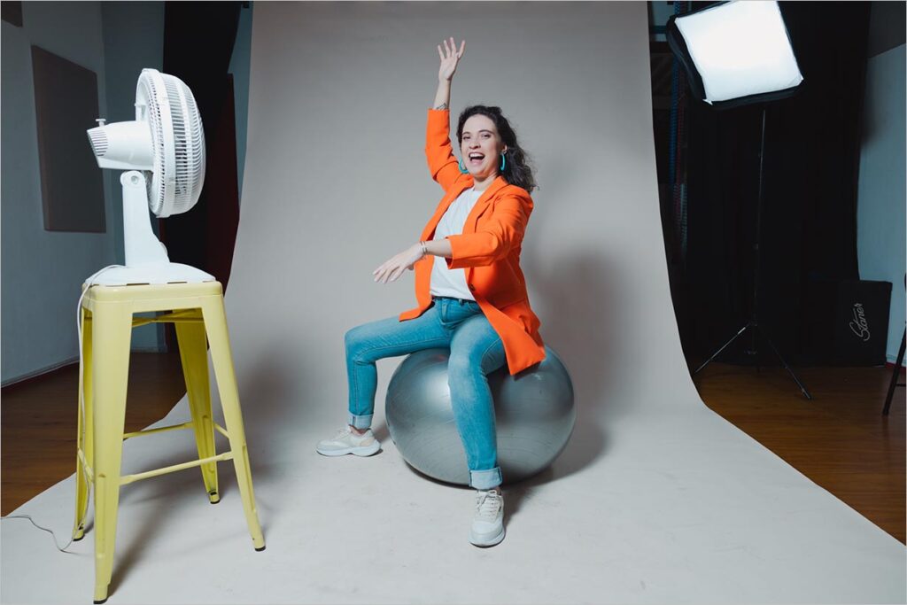 Mulher com roupas coloridas está sentada em uma bola prateada de ginástica e sorri levantando um dos braços. Do seu lado esquerdo, um ventilador sobre um banco, está voltado para ela.