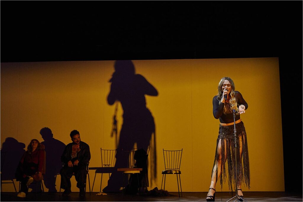 Uma cantora se apresenta ao microfone no palco, projetando uma grande sombra em uma parede iluminada de amarelo. Duas pessoas sentadas em cadeiras ao fundo assistem à apresentação. Foto: Nana Moraes