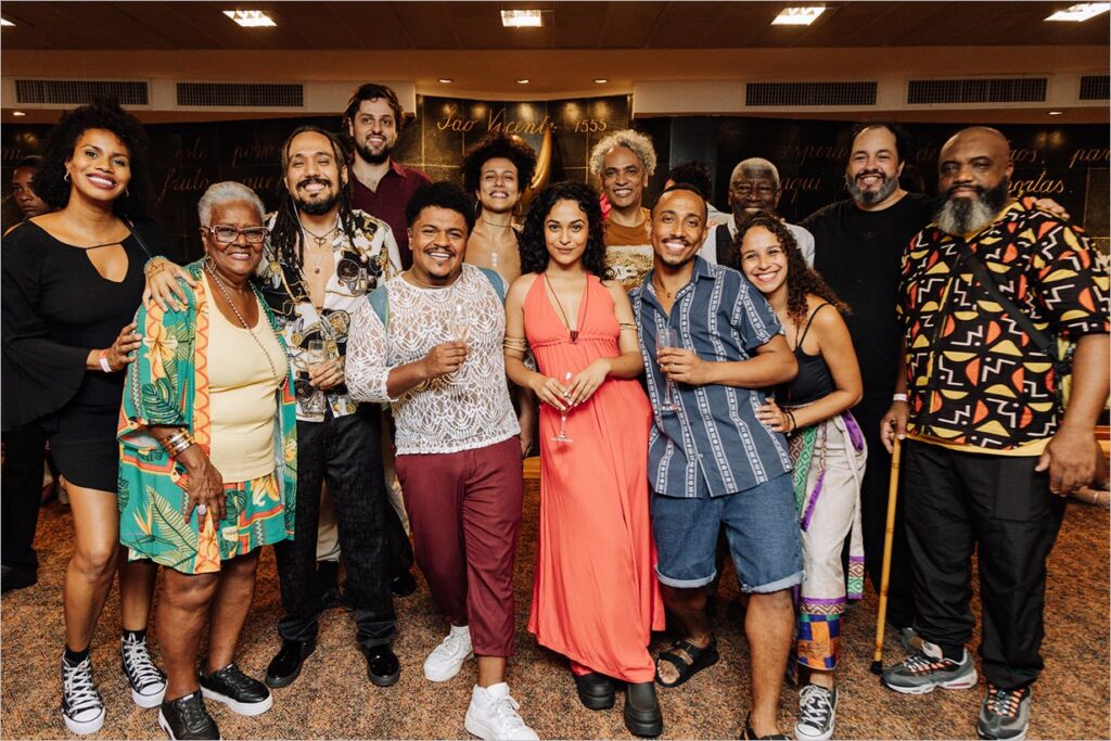 Um grupo diverso de pessoas, algumas sorrindo e outras segurando bebidas, posa junto em um ambiente interno. Uma pessoa usa uma bengala. A figura central usa um vestido coral, e o grupo está próximo, sugerindo uma celebração ou reunião social. Foto: Noelia Nájera
