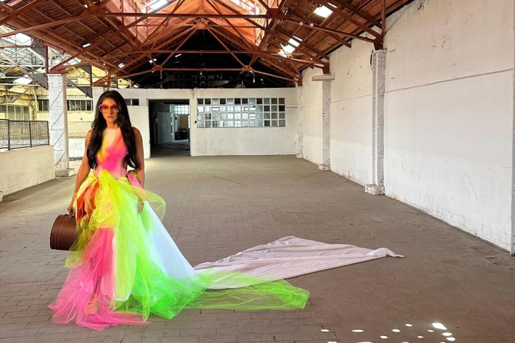 Uma mulher com um vestido multicolorido e brilhante, com cauda longa, está em um armazém industrial vazio. A luz do sol se filtra pelo teto, projetando sombras no chão. Ela segura uma mala marrom e usa óculos escuros. Foto: Karla Tenório