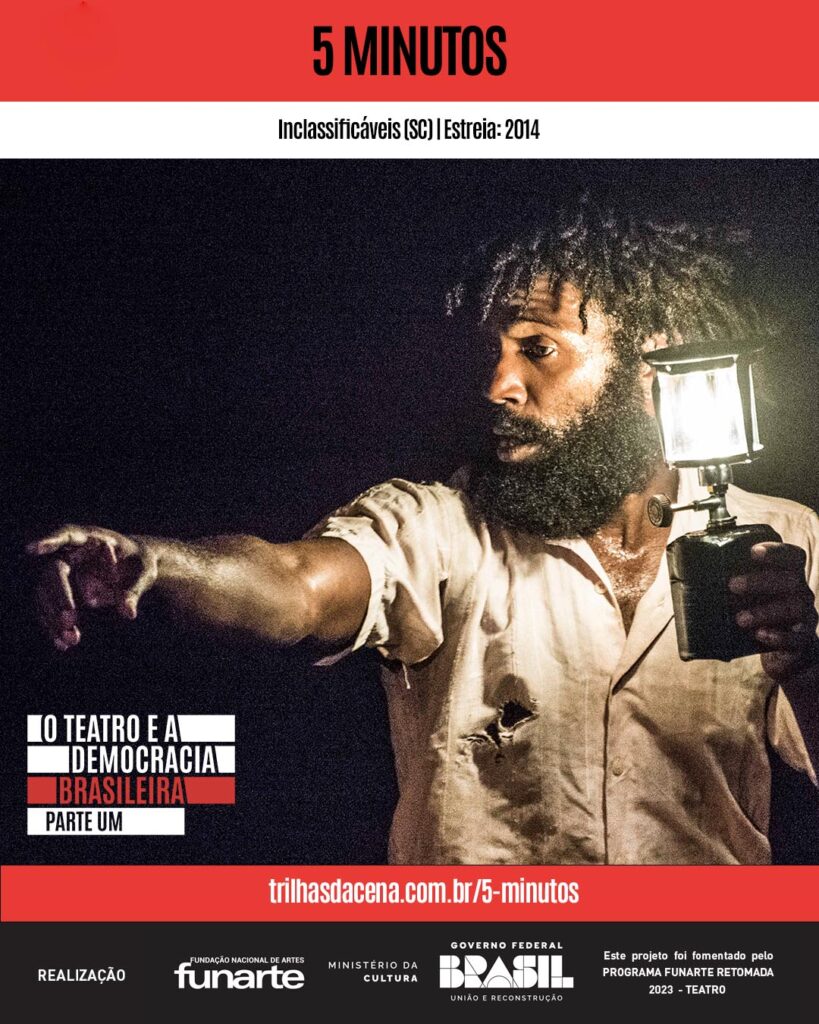 Um homem negro com barba segura uma lanterna, iluminando seu rosto e parte superior do corpo na escuridão. Ele está vestindo uma camisa de cor clara com pequenos rasgos, e aponta para a distância com a outra mão. O texto traz informações sobre o endereço eletrônico da página do espetáculo 5 minutos no Trilhas da Cena. Foto: Rodrigo Sambaqui