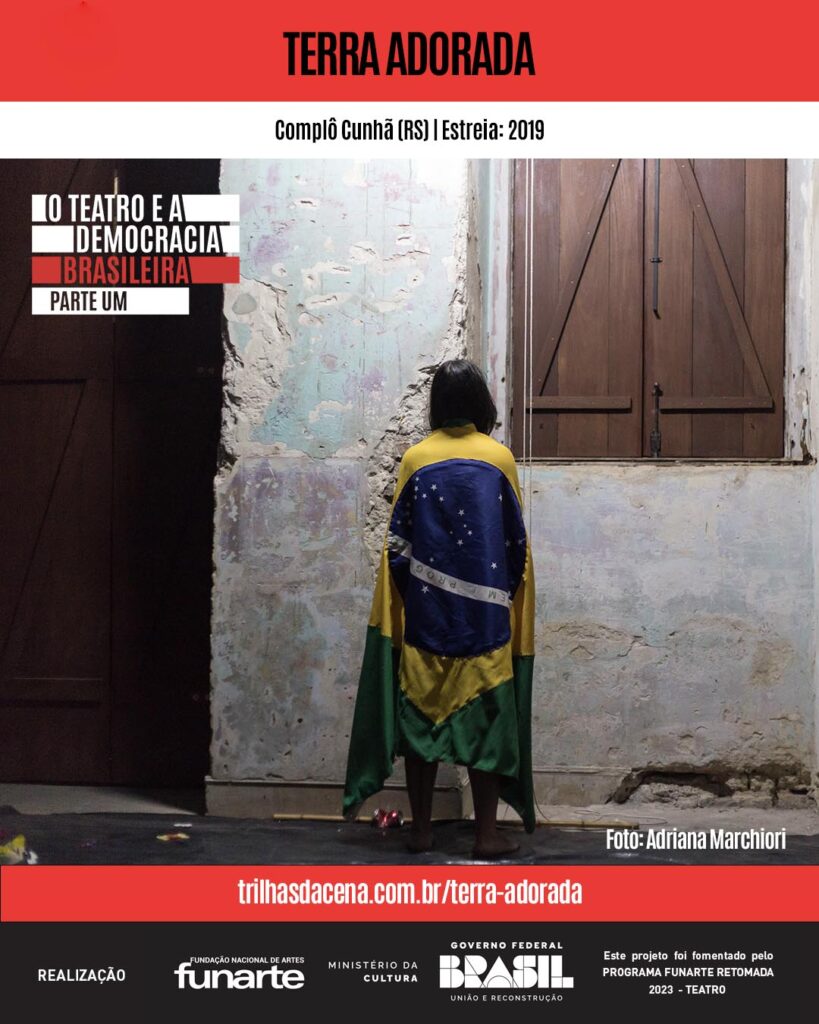 Uma mulher está descalça, de frente para uma parede, envolta em uma bandeira brasileira, com uma janela de madeira à direita. Acima, está o nome do espetáculo Terra adorada. A foto é de Adriana Marchiori
