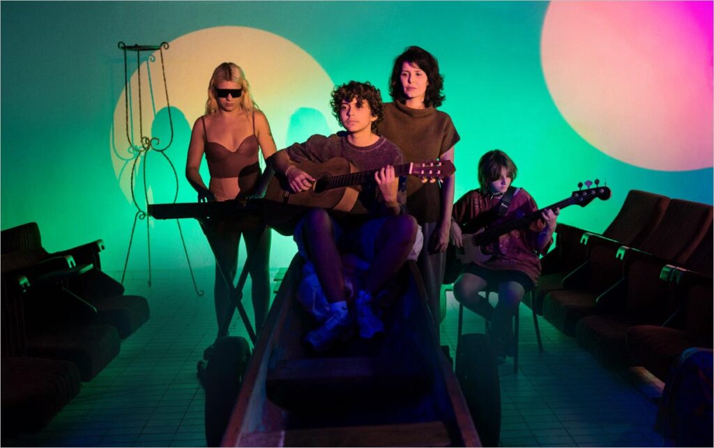 Quatro pessoas com instrumentos musicais sentam-se e ficam de pé em uma sala pouco iluminada com luzes coloridas e circulares na parede. Uma pessoa toca violão, outra toca baixo e uma terceira toca teclado, enquanto a quarta está no meio deles e observa. Foto: Ana Alexandrino