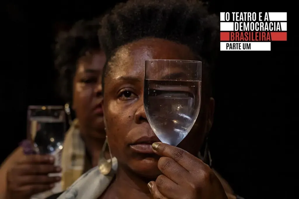 Uma mulher segura um copo de água na frente do rosto, distorcendo seu olho através do vidro. Outra mulher está atrás dela. O texto diz: O TEATRO E A DEMOCRACIA BRASILEIRA PARTE UM.