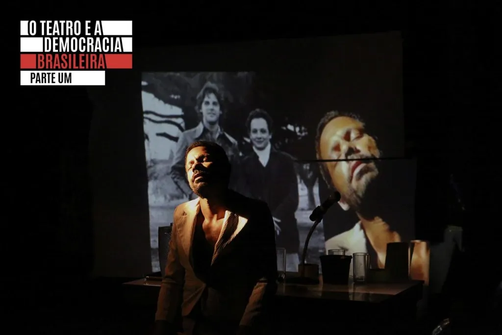 Um homem está em pé sob uma iluminação dramática diante de uma imagem projetada em preto e branco de três pessoas. No canto superior esquerdo, o texto diz: O TEATRO E A DEMOCRACIA BRASILEIRA PARTE UM.