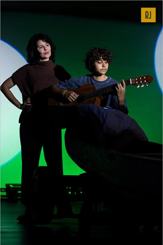 Um garoto com cabelo curto e cacheado toca violão enquanto está sentado. uma mulher com cabelo de comprimento médio está de pé ao lado dele com uma mão no quadril. Eles estão na frente de um fundo verde e azul com formas circulares. Foto: Ana Alexandrino