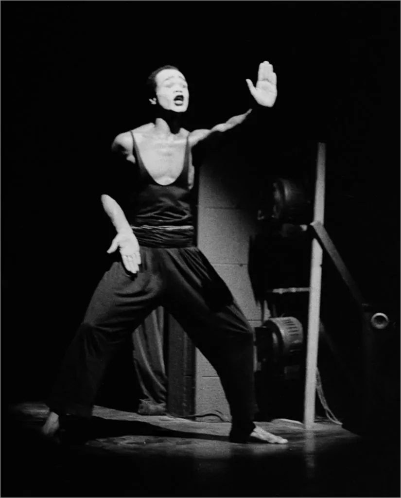 Uma figura se apresenta no palco, vestindo uma roupa escura sem mangas. O artista está com a boca aberta e uma mão levantada em um gesto expressivo. Holofotes apagados do palco são visíveis no fundo, projetando sombras dramáticas. Foto: Guto Muniz