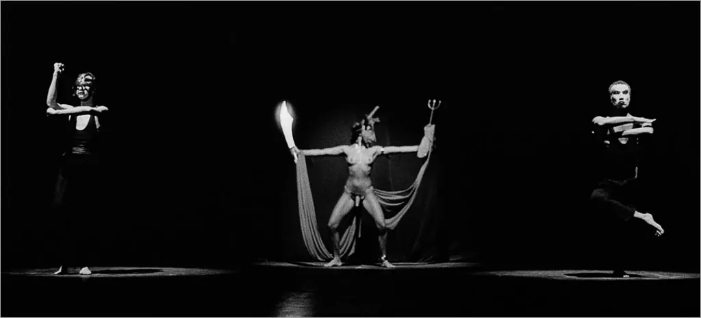 Uma performance dramática em preto e branco com três artistas. A artista central está usando uma máscara e um traje esvoaçante, estendendo os braços graciosamente parcialmente de pé. Os dois artistas laterais são pegos no meio do movimento, com braços e pernas equilibrados. O palco está com uma iluminação suave e focada nas figuras. Foto: Guto Muniz