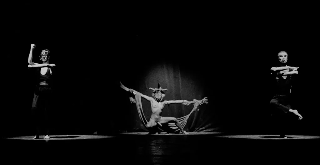 Uma performance dramática em preto e branco com três artistas. A artista central, agachada com uma perna parcialmente estendida, está usando uma máscara e um traje esvoaçante, estendendo os braços graciosamente. Os dois artistas laterais são pegos no meio do movimento, com braços e pernas equilibrados. O palco está com uma iluminação suave e focada nas figuras. Foto: Guto Muniz