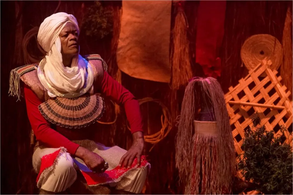 Um homem em traje tradicional está sentado, usando um turbante branco e uma peça de peito ornamentada, como o traje de um guerreiro africano. O cenário apresenta materiais texturizados e tons terrosos, com elementos tecidos e vegetais. Há um objeto que lembra uma pequena escultura ao lado dela. Foto: Guto Muniz