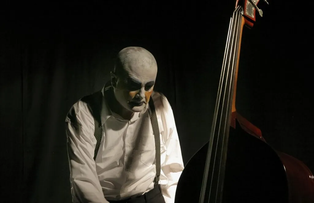 Um homem com pintura facial branca e detalhes escuros, como um mimico, vestindo uma camisa branca com suspensórios, toca um contrabaixo em um palco, sob um holofote suave. O fundo é escuro. Foto: Guto Muniz