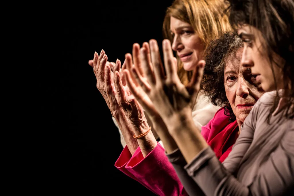 Três mulheres com expressões variadas estão em fila, parcialmente visíveis no canto direito da imagem, com as mãos levantadas na altura de seus rostos em foco. Elas estão vestidas em tons de vermelho, branco e cinza, em um fundo escuro. Foto: Renato Mangolin
