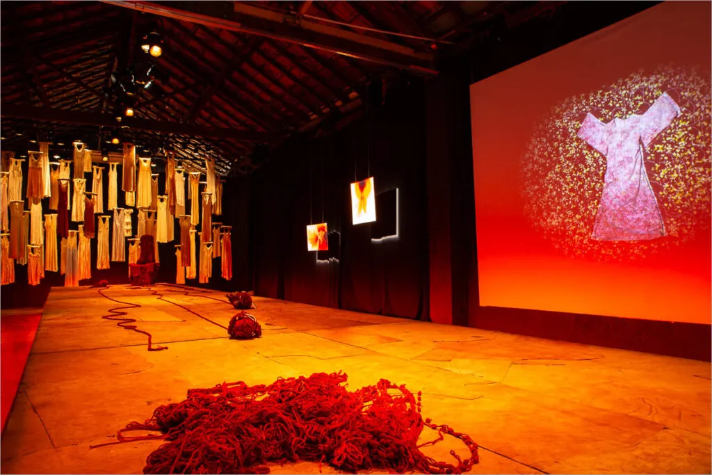 Uma sala iluminada por uma luz alaranjada e dispersa apresenta um palco, como uma passarela, com vestidos suspensos pelos cabides ao fundo e iluminação avermelhada. Uma tela grande, na lateral do palco, exibe a imagem de um vestido. Pilhas de um fio vermelho repousam no chão de maneira desorganizada. Foto: Renato Mangolin