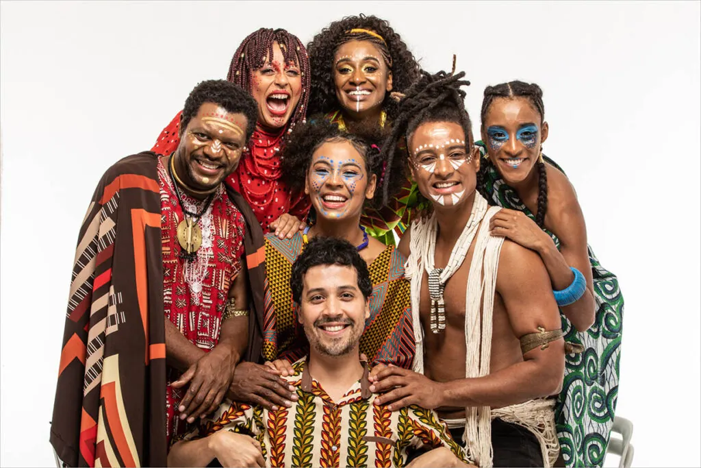Um grupo diverso de sete pessoas vestindo roupas vibrantes e tradicionais africanas posa junto, sorrindo. Eles têm pintura facial colorida e estão posicionados contra um fundo branco simples. Foto: Renato Mangolin