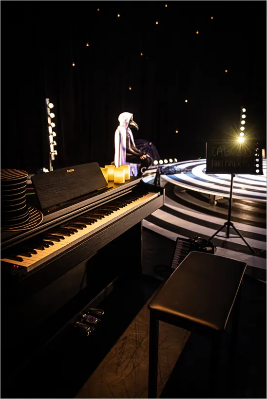 Um palco pouco iluminado apresenta um teclado e um banco em primeiro plano. No fundo, um artista senta-se em uma plataforma circular listrada, vestido de uma capa branca com capuz e uma máscara dos doutores da peste negra. Pequenas luzes pontilham o cenário, e uma placa com uma frase pouco visível está em cena. Foto: Renato Mangolin