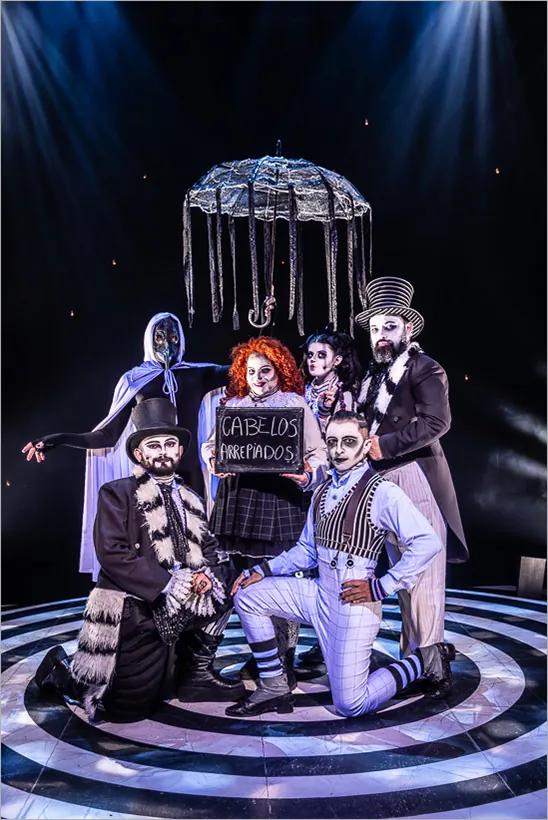 Seis artistas, homens e mulheres, em trajes teatrais e maquiagem de caveiras preta e branca posam em um palco de desenho espiral. Um segura um pequeno quadro negro dizendo "Cabelos Arrepiados". Outro segura uma sombrinha de tecido com fitas de tecido preto pendurados nas beiradas O fundo é escuro com luzes parecidas com estrelas, e um holofote brilha de cima. Foto: Renato Mangolin