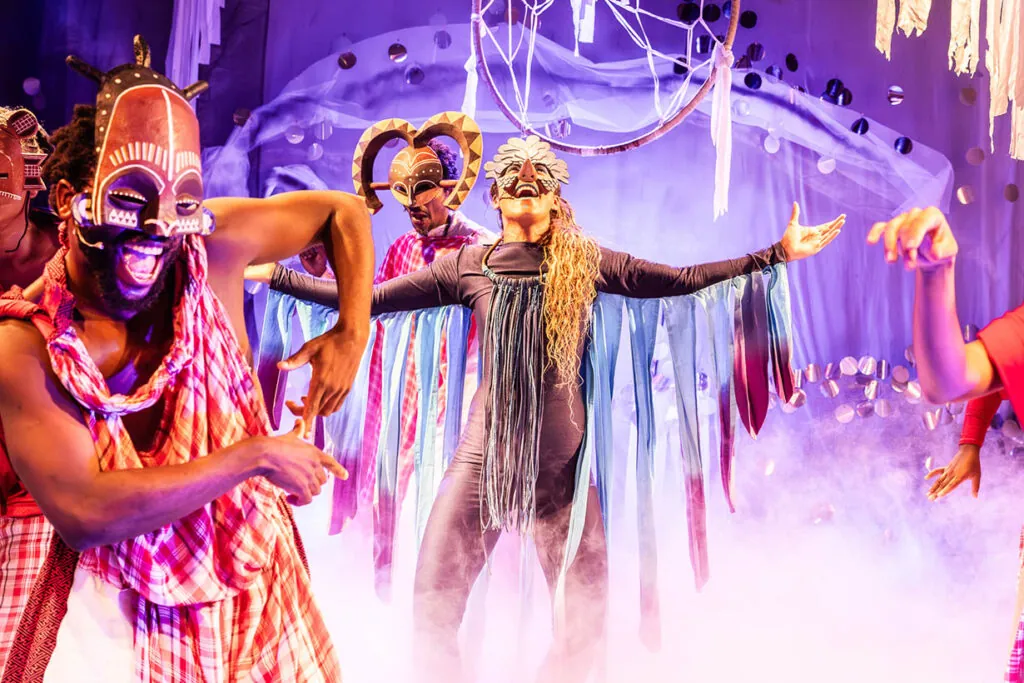 Artistas em máscaras africanas e trajes tradicionais e coloridos dançam empolgadamente em um palco enevoado iluminado com vibrantes roxos e azuis. Eles usam fitas esvoaçantes e elaborados adereços de cabeça, criando uma atmosfera energética e festiva. Foto: Renato Mangolin