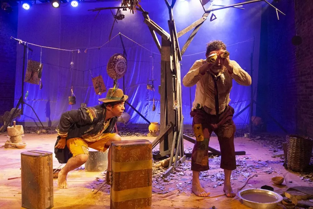 Dois atores atuam no palco. Um agacha-se com uma roupa colorida e um chapéu, enquanto o outro fica de pé, usando um binóculo improvisado e roupas esfarrapadas. O cenário inclui uma árvore improvisada e adereços espalhados ao redor. A iluminação é dramática e temperamental. Foto: Guto Muniz