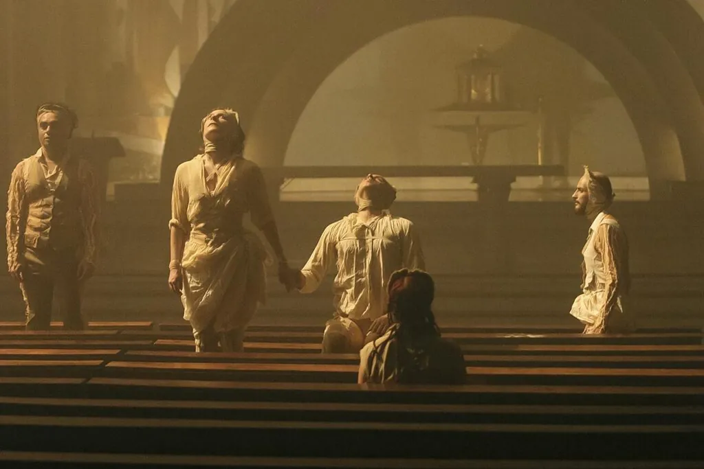 Quatro pessoas em trajes teatrais estão dramaticamente de pé com os braços ao lado do corpo entre bancos de igreja, de frente para direções diferentes. Outra pessoa está sentada de costas para o espectador, olhando para elas. O cenário tem uma iluminação atmosférica e dourada. Foto: Guto Muniz