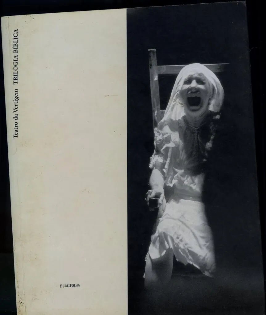 Uma imagem em preto e branco captura uma pessoa com uma expressão aflita e boca aberta, possivelmente gritando ou berrando. Vestida de branco com um lenço na cabeça, ela está parcialmente na sombra. O texto à esquerda diz Teatro da Vertigem TRILOGIA BÍBLICA, destacando um espetáculo do Teatro da Vertigem.