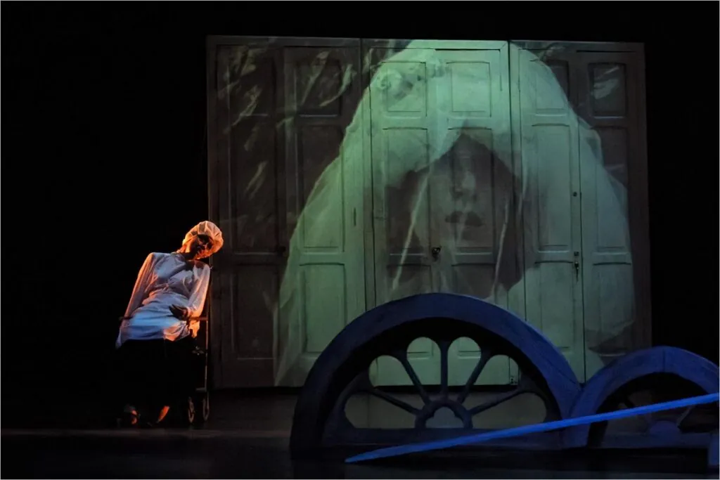 Uma pessoa com roupa branca e boné está sentada em uma cadeira sob iluminação fraca. Uma grande projeção do rosto de uma noiva com um véu aparece em um fundo de portas com painéis. No primeiro plano, duas grandes formas de rodas são parcialmente visíveis. Foto: Guto Muniz