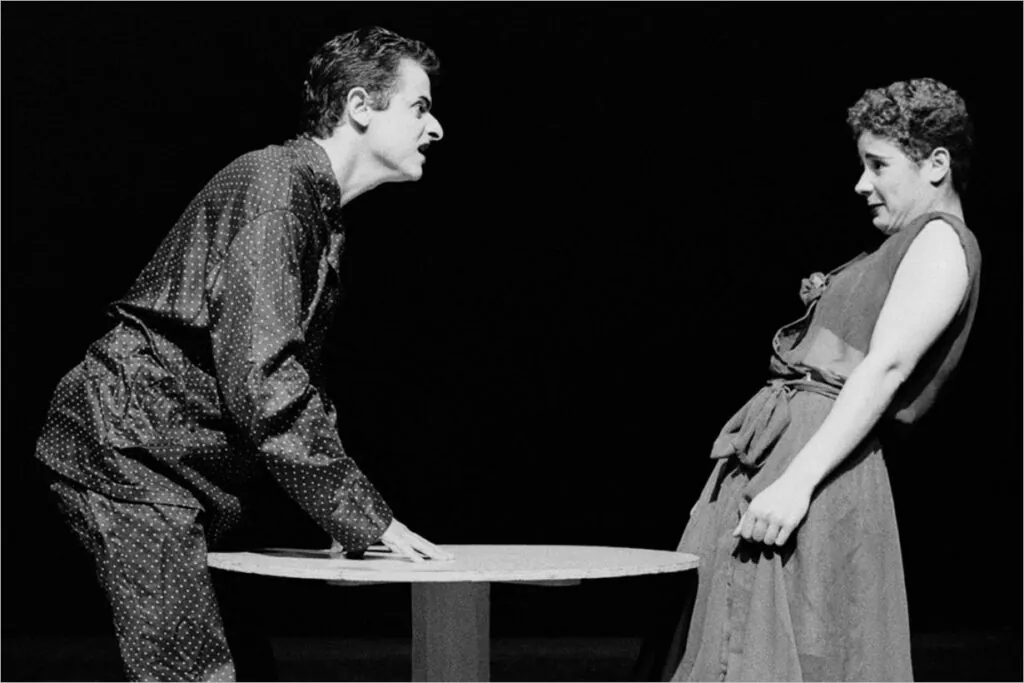 Uma imagem em preto e branco de dois atores atuando no palco. Um homem, vestido com um pijama, está inclinado sobre uma mesa, olhando intensamente, enquanto uma mulher se inclina para trás com uma expressão de apreensão. Foto: Guto Muniz