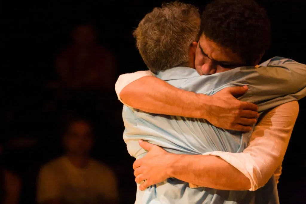 Dois homens estão se abraçando fortemente à direita da imagem, um usando uma camisa azul-bebê e o outro usando uma camisa branca. O fundo é escuro, e algumas pessoas estão borradas nas sombras. A iluminação é suave e quente. Foto: Guto Muniz