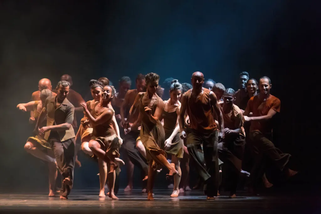 Um grupo de dançarinos e dançarinas se apresentando energicamente no palco. Eles estão usando trajes em tons de terra, e a iluminação cria uma atmosfera dramática com tons escuros e melancólicos. Os dançarinos estão se movendo em uníssono com poses dinâmicas e expressivas. Duas dançarinas no palco fazem uma pose dramática. A dançarina da esquerda está de cabeça para baixo, usando uma saia vermelha que cobre seu tronco de cabeça pra baixo, parecida com uma rosa. O dançarino da direita se inclina para frente em uma roupa preta e dourada. O fundo é escuro e somente as dançarinas são sutilmente iluminadas. Foto: Guto Muniz