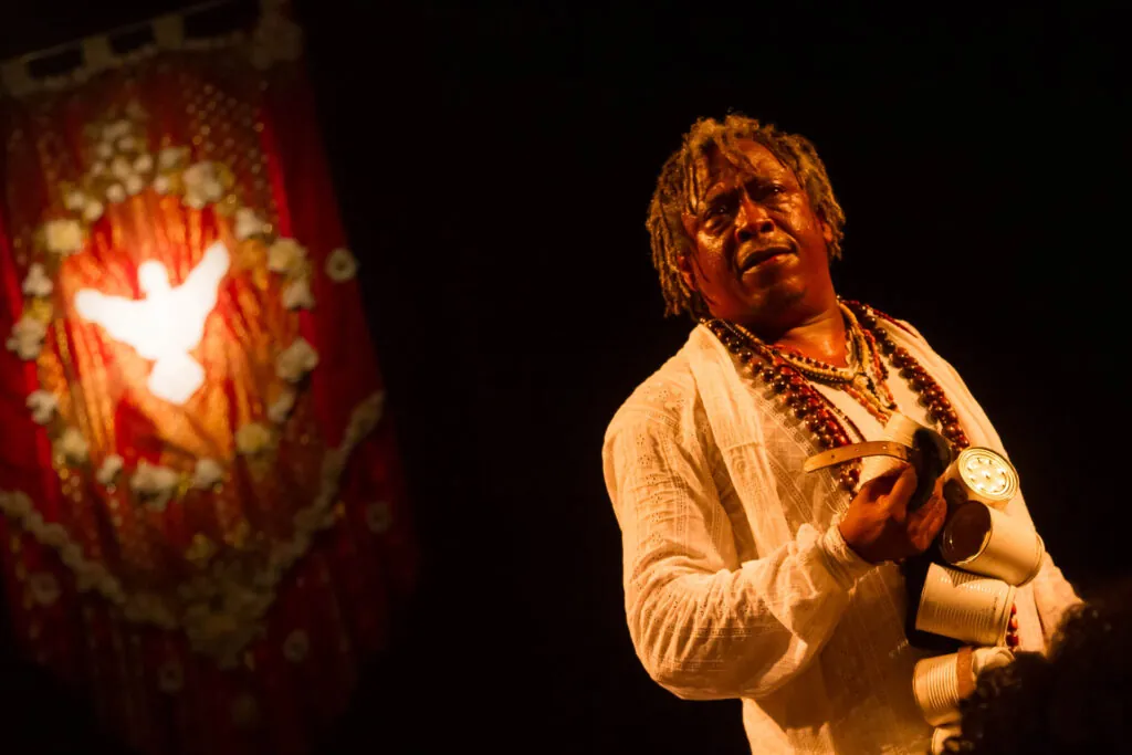 Um homem vestindo uma camisa branca e colares de contas segura algumas latinhas de metal, pintadas de branco, em sua mão esquerda. Ele olha para cima com uma expressão de emoção. Uma faixa vermelha com um desenho de pomba branca é visível ao fundo. Foto: Guto Muniz