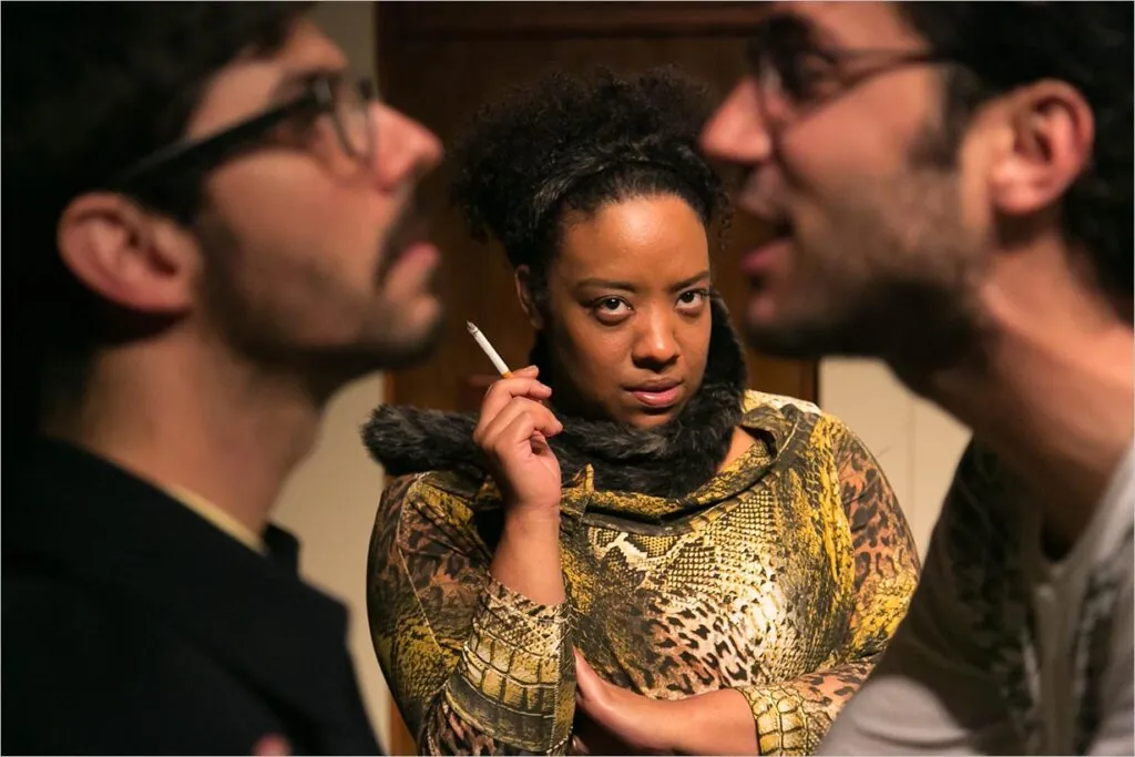 Uma mulher com uma expressão focada e desdenhosa segura um cigarro, enquanto observa dois homens parcialmente visíveis em primeiro plano, desfocados, emoldurando seu rosto. Ela está usando uma roupa estampada e um cachecol de pele. O fundo é suavemente iluminado. Foto: Guto Muniz