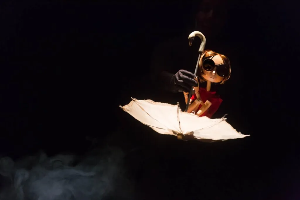 Uma boneca de madeira com cabelo curto e óculos redondos está sentada dentro de um guarda-chuva branco aberto. Uma mão usando uma luva preta segura o pequeno guarda-chuva por trás. A cena é suavemente iluminada contra um fundo escuro. Foto: Guto Muniz