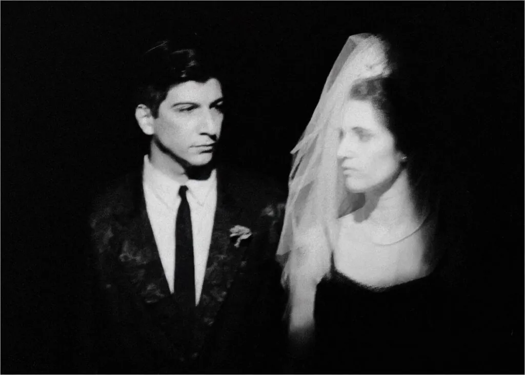 Uma imagem em preto e branco de um homem de terno e gravata em pé ao lado de uma mulher em um vestido e um véu cobrindo seu resto. Eles estão colocados contra um fundo escuro, ambos aparecendo em um foco suave. Foto: Guto Muniz