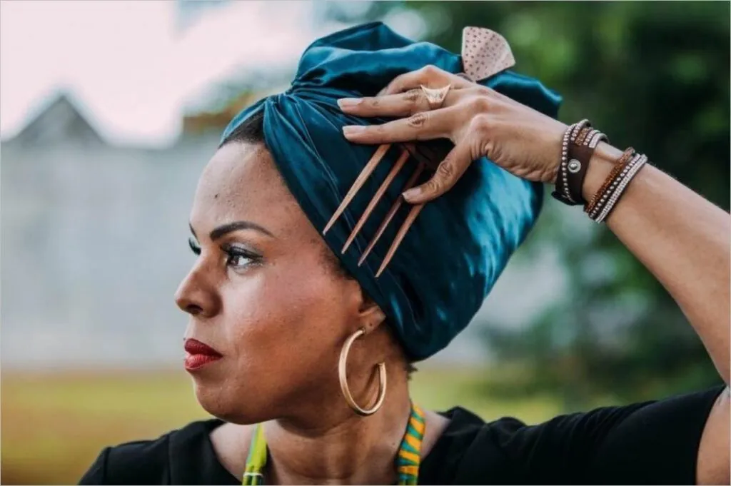 Uma mulher negra usando um turbante azul e grandes brincos de argola olha para o lado. Ela levanta a mão para ajustar o turbante, com longas unhas vermelhas e várias pulseiras no pulso. O fundo é desfocado com vegetação e um muro. Foto: Thaís Mallon