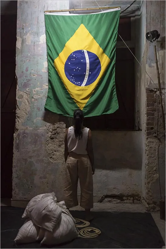 Uma mulher está de costas, contemplando uma grande bandeira brasileira em uma parede desgastada. A parede tem tijolos expostos e tinta descascada, com sacos e cordas enroladas espalhadas no chão ao lado. Foto: Adriana Marchiori