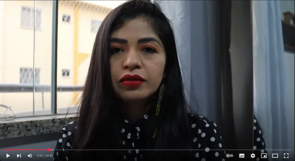 Mulher de longos cabelos escuros e batom vermelho sentada em um ambiente interno, vestindo uma camisa de bolinhas. Ao fundo, uma janela com vista para um prédio. Barra de progresso do vídeo visível na parte inferior.