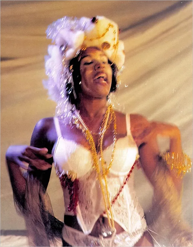 Uma mulher negra dança e canta vestida com um traje branco e um adorno de cabeça com penas e flores. Ela usa colares em camadas e pulseiras douradas, criando uma atmosfera festiva e vibrante contra um fundo drapeado. Foto: Luiz Alves