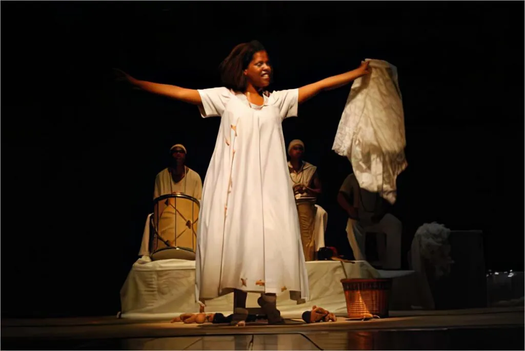 Uma mulher negra no palco veste um vestido branco, sorri com os braços estendidos e segura um pano. Dois músicos em trajes semelhantes sentam-se ao fundo com um tambor. Cestos e panos estão no chão. Foto: Luiz Alves