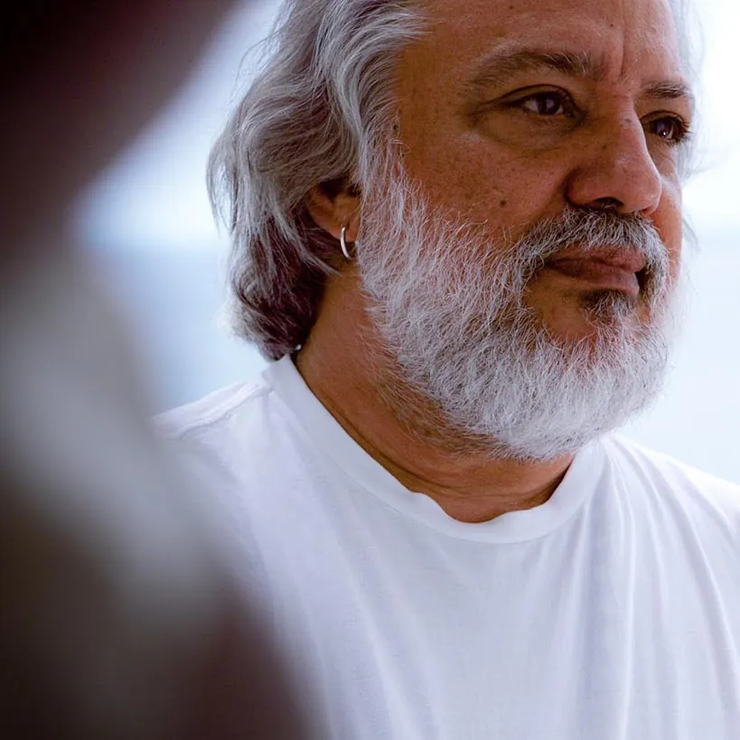 Um homem de cabelos grisalhos e barba branca, vestindo uma camisa branca, olha à distância, em pé, ao ar livre, com um fundo desfocado. Foto João Milet Meirelles