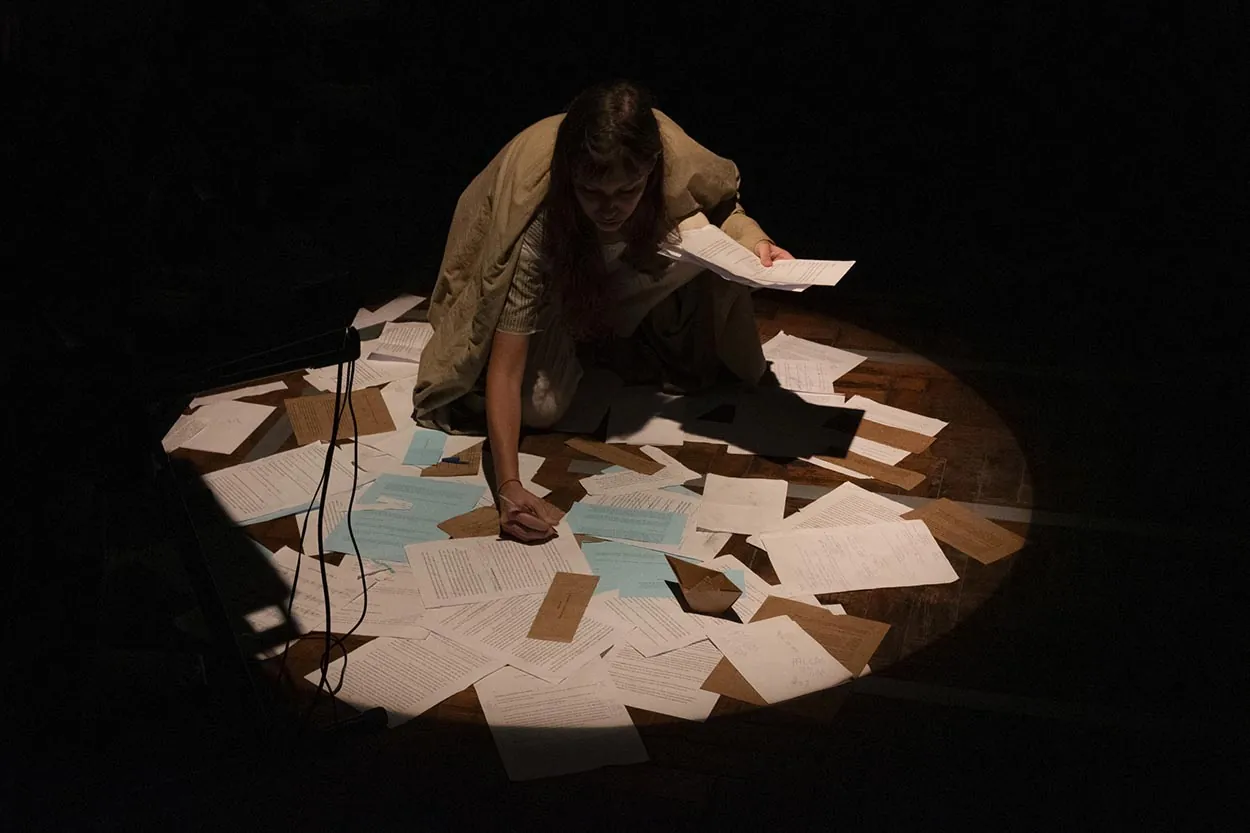Uma mulher se ajoelha sob um holofote em um palco pouco iluminado, cercada por papéis espalhados. Ela está segurando alguns papéis e escreve eu um que está no chão. Foto: Jorge Alves