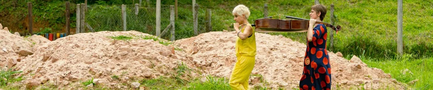 Duas mulheres caminhando na grama perto de montes de areia. Uma usa um macacão amarelo e a outra usa um vestido escuro com círculos vermelhos. Elas seguram um mesmo violoncelo nos ombros. Árvores e uma cerca estão ao fundo. Foto: Thaís Grechi