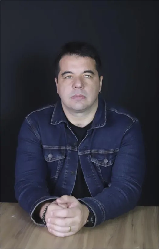 Homem sentado em uma mesa de madeira, olhando diretamente para a câmera com uma expressão séria. Ele veste uma jaqueta jeans azul sobre uma camisa preta e tem as mãos cruzadas à frente do corpo. O fundo é preto, destacando o rosto e a postura dele. Foto: João Pedro Ribeiro