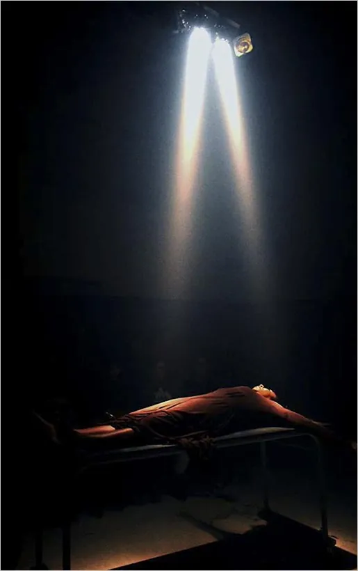 Uma pessoa está deitada em uma mesa sob feixes de luz focados e dramáticos em um espaço escuro, criando uma atmosfera teatral e misteriosa. Foto: Regina Peduzzi Protskof